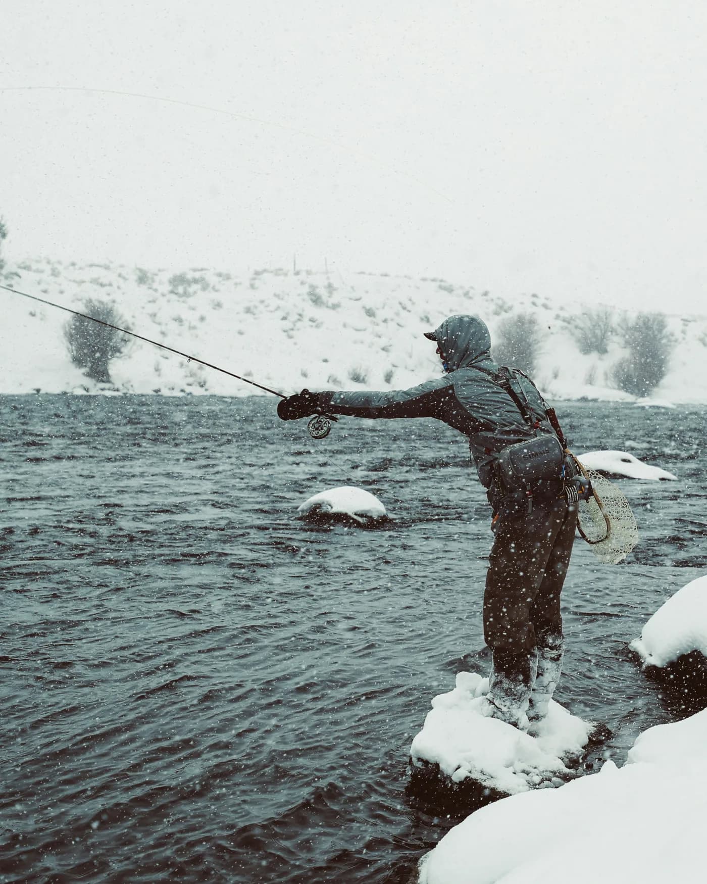 An angler in waders stands on a snow-covered rock, casting a fly rod across a dark river during a snowstorm.